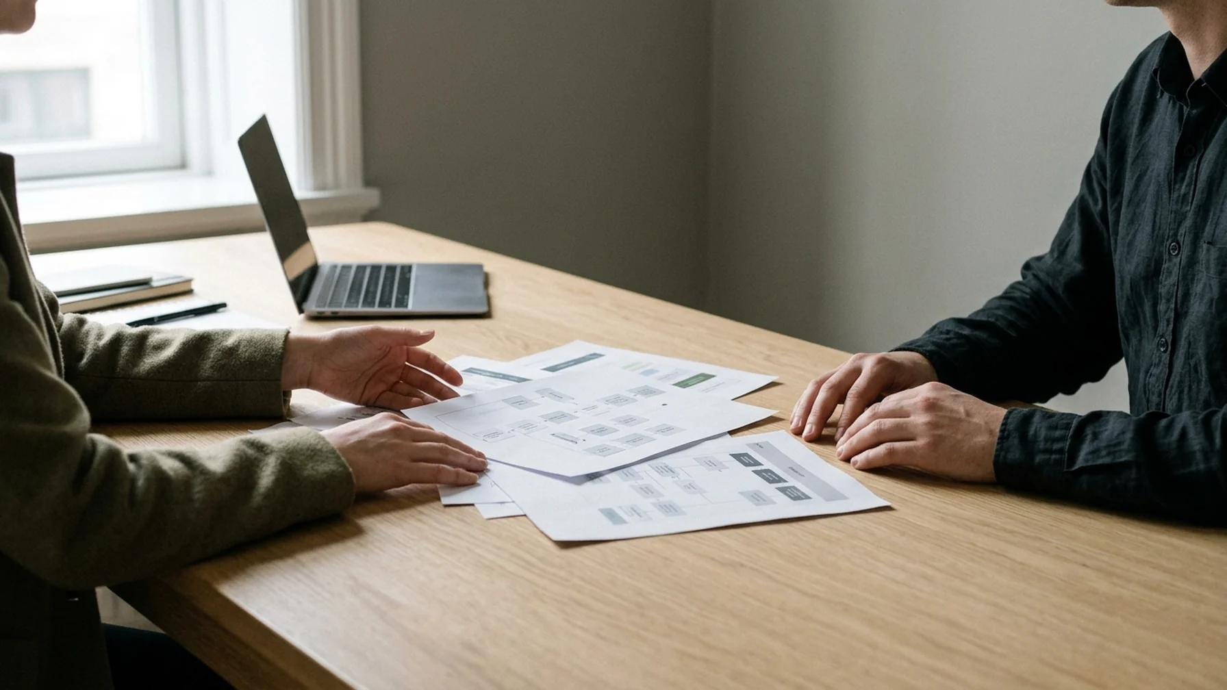 A small business owner and a consultant at a wooden table reviewing printed process diagrams beside a laptop, with daylight from a window across the papers.